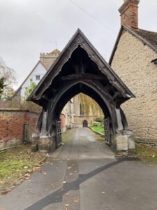 A wooden lychgate with a steep roof stands at the entrance to a churchyard along the Thames Path, flanked by old brick and stone buildings, with a pathway leading to a church in the background.