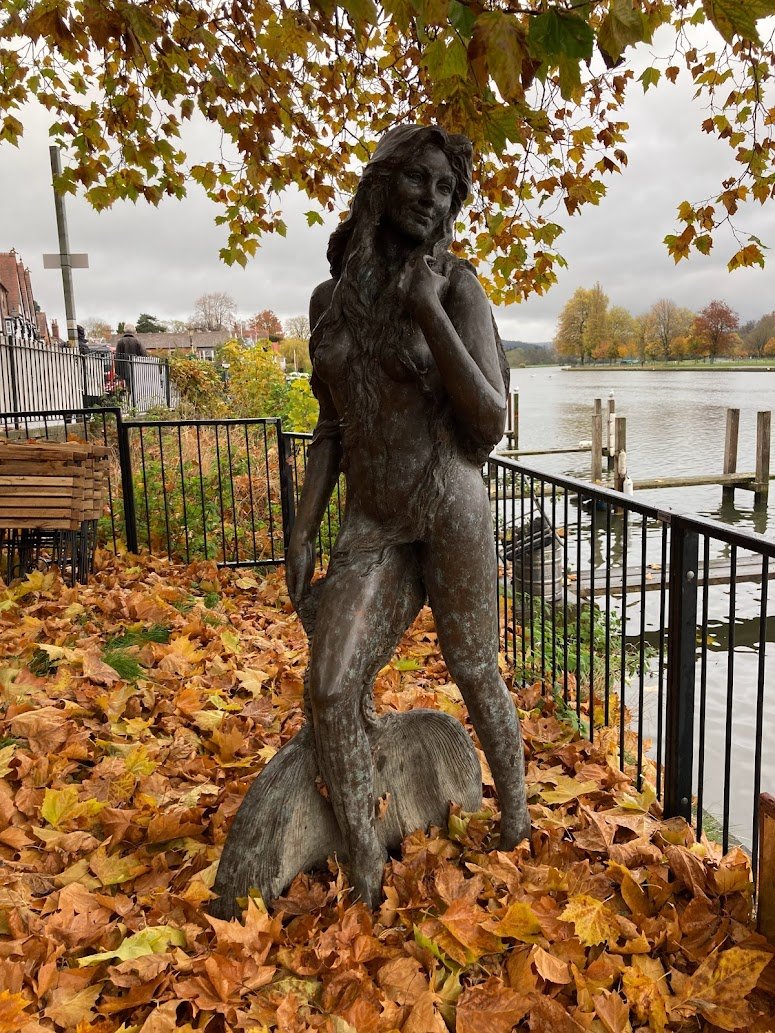A bronze statue of a woman with long hair stands on fallen autumn leaves near a river, with trees, fencing, and docks visible in the background under a cloudy sky.
