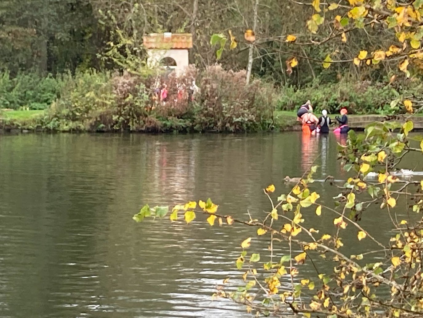 People in bright clothing stand at the edge of a calm lake near dense greenery and a small decorative stone structure; yellow leaves are visible in the foreground.