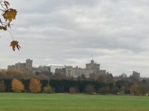 A distant view of a large stone castle with a central round tower and battlements, surrounded by autumn trees and a grassy field under a cloudy sky.