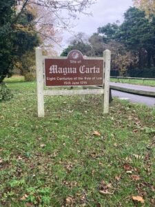 A brown sign on grass reads: Site of Magna Carta. Eight Centuries of the Rule of Law. 15th June 1215. Trees and the nearby Thames Path are in the background.