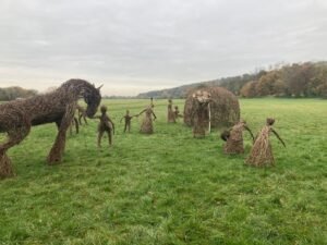 Large woven willow sculptures of people, a horse, and an elephant stand playfully on a grassy field along the Thames Path under a cloudy sky, with trees in the background.