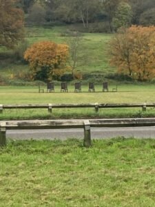 Five wooden chairs are lined up in a grassy field with autumn trees in the background; a wooden fence and a road, reminiscent of the Thames Path, run across the foreground.