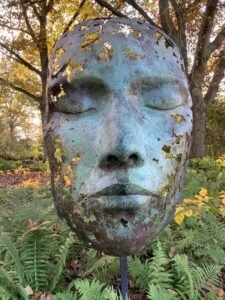 A large, weathered metal sculpture of a serene human face with closed eyes stands among green ferns and autumn trees. Parts of the mask are missing, revealing holes and jagged edges.