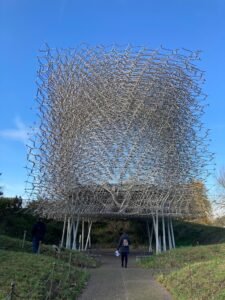 A person walks toward a large, intricate metal sculpture resembling a beehive, elevated on stilts, set outdoors under a clear blue sky with grassy areas and trees nearby.