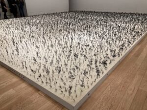 A large, square white platform covered densely with small, upright metal sculptures resembling abstract plants or flowers, displayed in a gallery with wooden floors and white walls. Several people stand in the background.