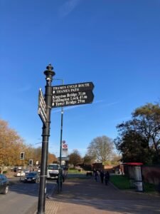 A black signpost shows directions for the Thames Cycle Route and Thames Path, pointing toward Kingston Bridge (3¼ mi), Teddington Lock (4½ mi), and Tower Bridge (29 mi) on a sunny day with a clear blue sky.