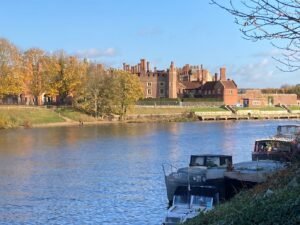 A large historic building with many chimneys sits across the river along the Thames Path, framed by autumn trees and a blue sky. Several boats are moored in the foreground along the riverbank.
