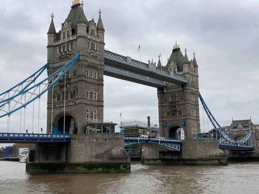 The image shows London’s iconic Tower Bridge, with its two tall towers and blue suspension elements spanning over the River Thames on a cloudy day.