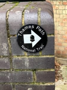 A round black-and-white sign says Thames Path National Trail with a white arrow pointing right, mounted on a wet, dark brick wall. Theres some green moss between the bricks. A red brick wall is in the background.
