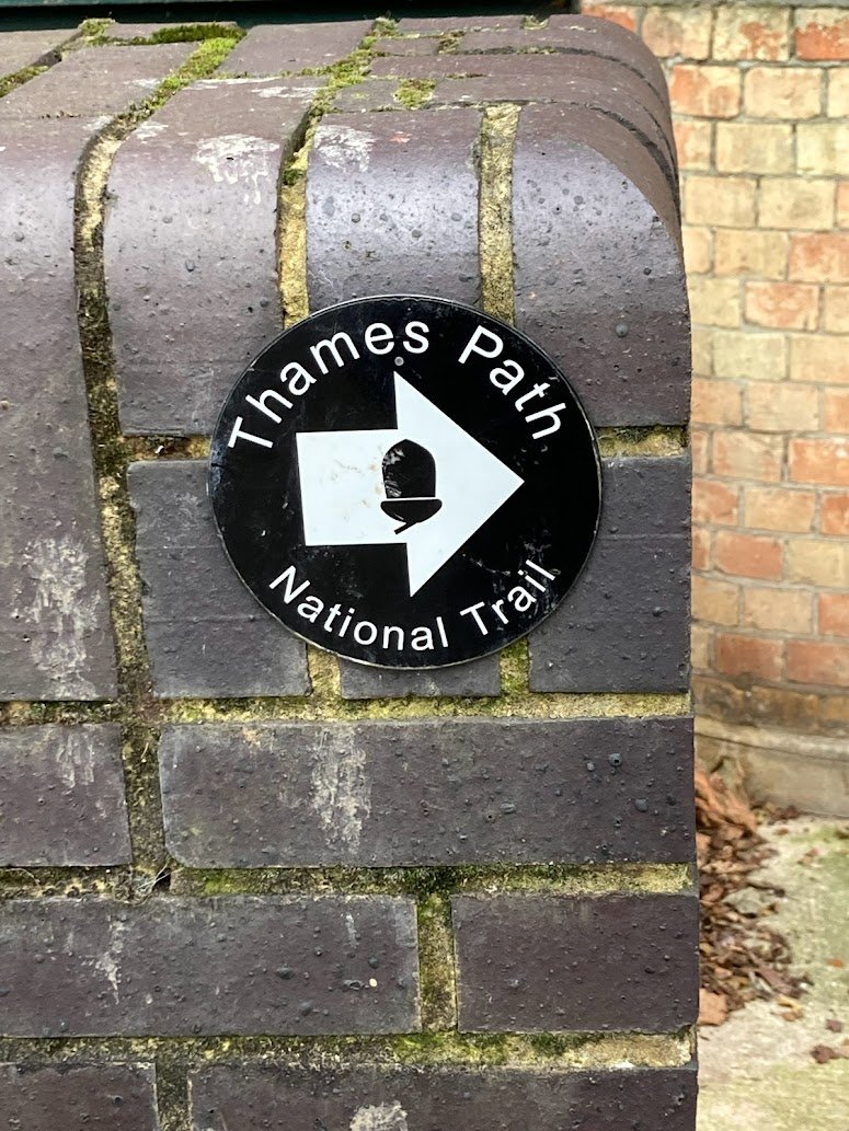 A round black-and-white sign says Thames Path National Trail with a white arrow pointing right, mounted on a wet, dark brick wall. Theres some green moss between the bricks. A red brick wall is in the background.