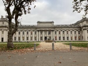 A large neoclassical building with columns and symmetrical windows, seen from across a lawn scattered with autumn leaves near the Thames Path. Several people are sitting or walking, and bare trees frame the scene under a cloudy sky.
