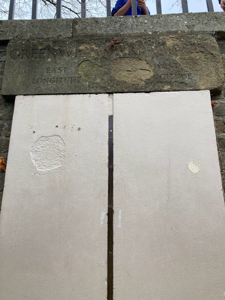A stone plaque above a wall marks the Greenwich Meridian Line, with faded text reading Greenwich Meridian and Longitude East/West. A vertical line runs down the wall, and a metal fence is visible at the top.