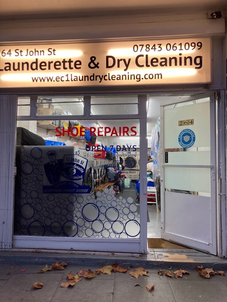 The entrance of a launderette and dry cleaning shop with a bright sign above the door, a shoe repairs sign, and autumn leaves scattered on the pavement outside. The shop window displays blue circular patterns and the open door shows laundry inside.