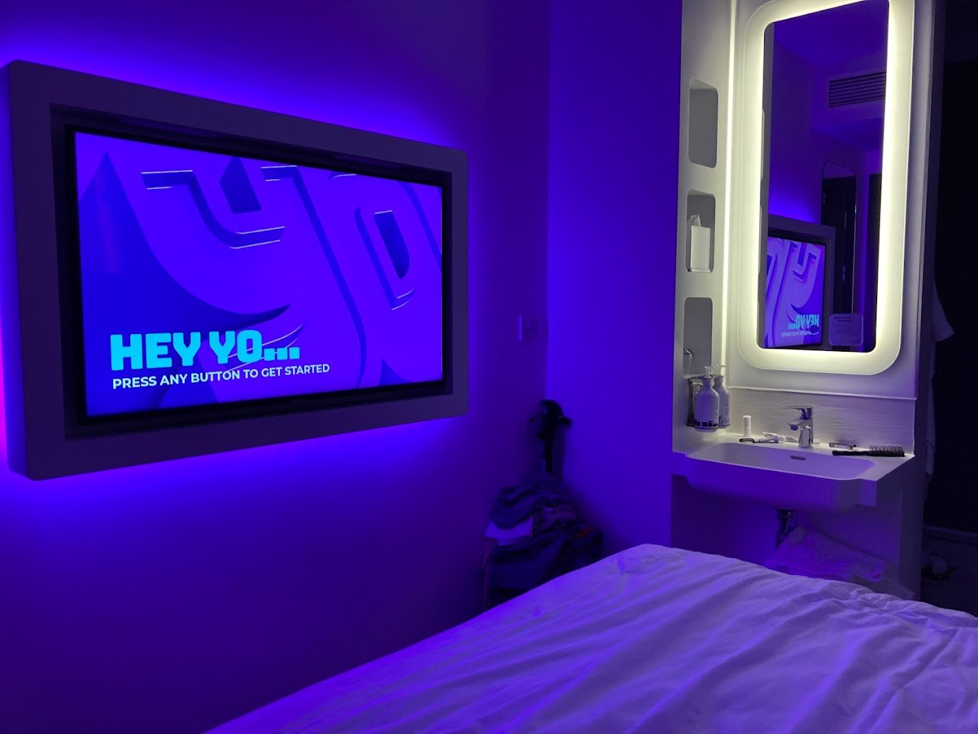 A modern hotel room with purple-blue lighting, showing a bed, a wall-mounted TV displaying HEY YO... PRESS ANY BUTTON TO GET STARTED, and a lit-up mirror above a small white sink with toiletries.