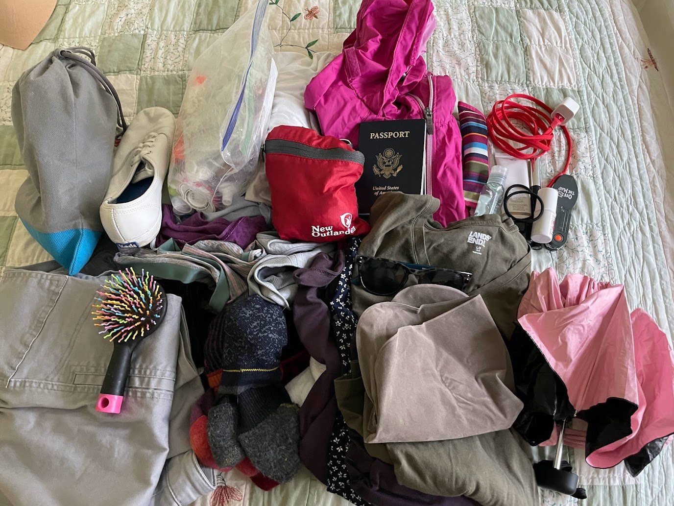 A variety of travel essentials, perfect for walking the Thames Path—including clothes, a passport, shoes, toiletries, an umbrella, a hairbrush, socks, a red cable, a rain jacket, and sunglasses—are spread out on a bed with a floral quilt.