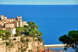 Sunlit buildings and lush green trees perch on a rocky cliff overlooking a calm, deep blue sea under a clear sky—a scene that perfectly captures the charm you'll discover on a day trip to Monaco. A small road or walkway runs along the waterfront below.